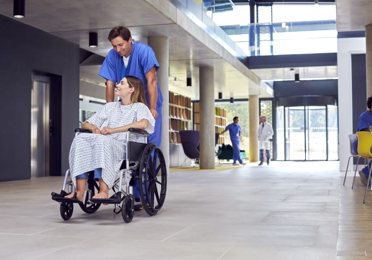 Male Nurse Wearing Scrubs Pushing Female Patient In Wheelchair Through Hospital Building